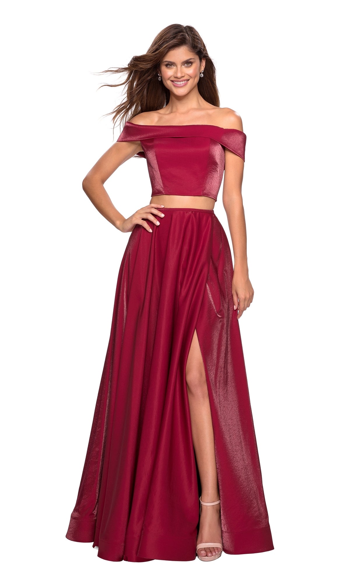 A woman poses, smiling in the La Femme A-Line Two-Piece Satin Prom Dress 26919—a burgundy off-the-shoulder crop top with a matching high-slit maxi skirt—showcasing its elegant a-line silhouette against a white background.
